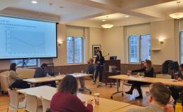 Xue Zhang, an Asian woman wearing a dark sweater, presents to the group in a wide shot of the room.