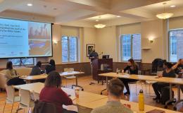 Nelson Roque, a white man wearing a blue shirt with a beard, presents to the group in a wide shot.