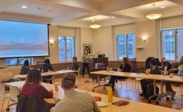 Tracee Saunders, a woman with shoulder-length brown hair wearing a green sweater, presents to the group in a wide shot. 