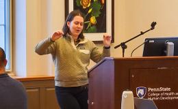 Tracee Saunders, a woman with shoulder-length brown hair wearing a green sweater, presents to the group. 