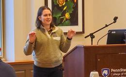 Tracee Saunders, a woman with shoulder-length brown hair wearing a green sweater, presents to the group. 