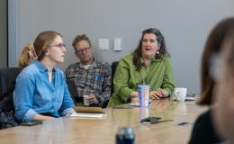 A woman in a green shirt talks to the group, sitting beside a man in a plaid shirt and a woman in a blue shirt.