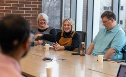 Three people sit at a conference table, a woman in a grey vest with white hair, a woman with blonde hair wearing brown overalls, and a man in a blue colored polo shirt.