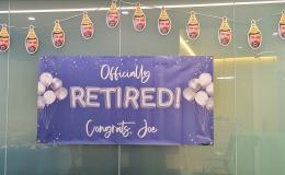 Blue sign that says "Officially Retired! Congrats Joe" on it with a banner of Joe's face hanging above it.