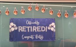 Blue sign that says "Officially Retired! Congrats Betsy" with a banner of Betsy's face hanging above it.