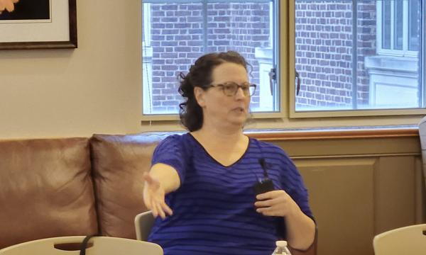 Stephanie Lanza, a white woman with glasses and brown hair in a blue sweater, talks to the group.