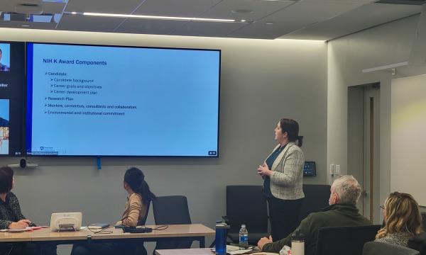 Cheryl Thompson, a woman wearing a white jacket, speaks to the group about NIH K awards.