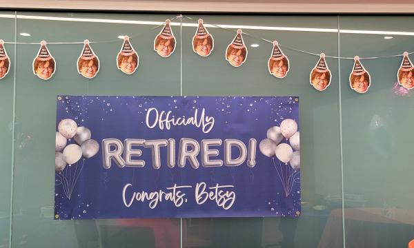 Blue sign that says "Officially Retired! Congrats Betsy" with a banner of Betsy's face hanging above it.